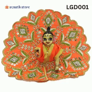 A small, brass Laddu Gopal (baby Krishna) deity idol is seated and dressed in a vibrant orange poshak (outfit). The outfit is heavily adorned with intricate gold embroidery, silver sequins arranged in diamond patterns, and green leaf-like designs. The deity has large, expressive eyes and a tilak on the forehead. The background is plain white, with a "Swastik Store" logo in the top left corner and the product code "LGD001" in the top right. - Swastik Store